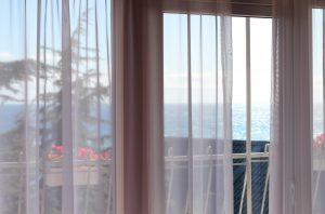 Looking through curtains at the coast | Featured image for the blog on Beachy Window Coverings from Cosmopolitan Shutters.
